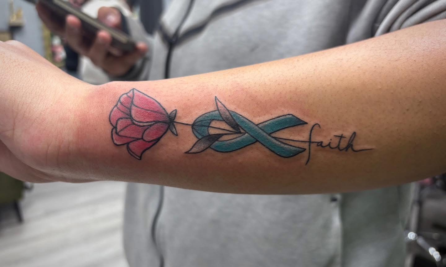 Teal ribbon, poppy and faith script tattoo on a forearm